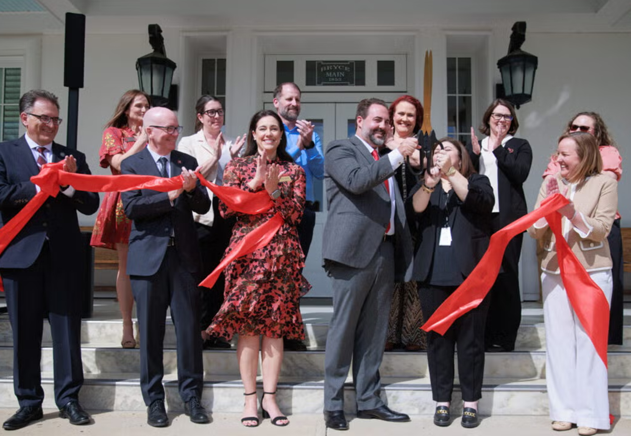 University leaders applaud as oversized scissors cut a crimson ribbon.