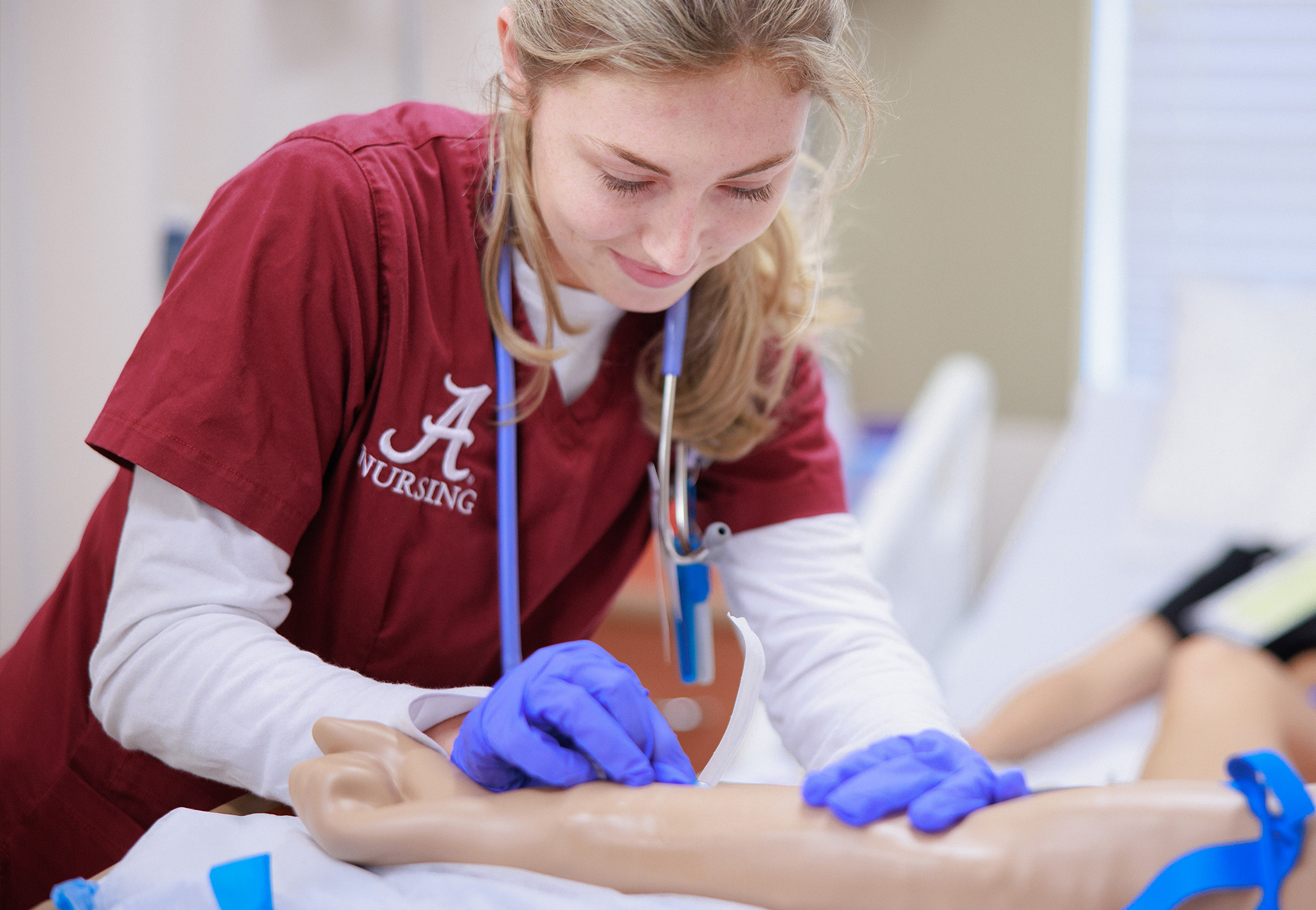 A nursing student in crimson scrubs and blue gloves practices starting an IV on a training manikin's arm