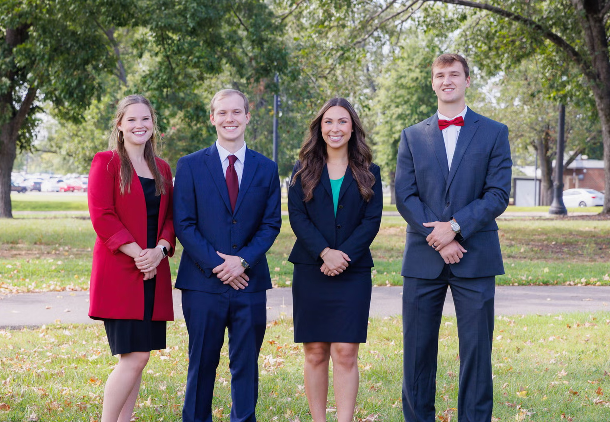 Four students in business attire stand together on a tree-lined lawn on campus