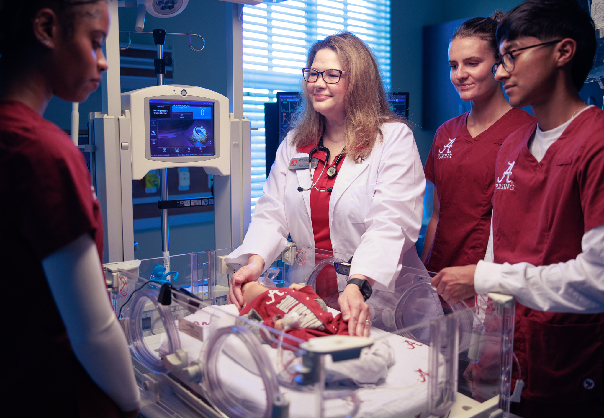 Dr. Dudding demonstrates infant care in an incubator to three students in crimson scrubs