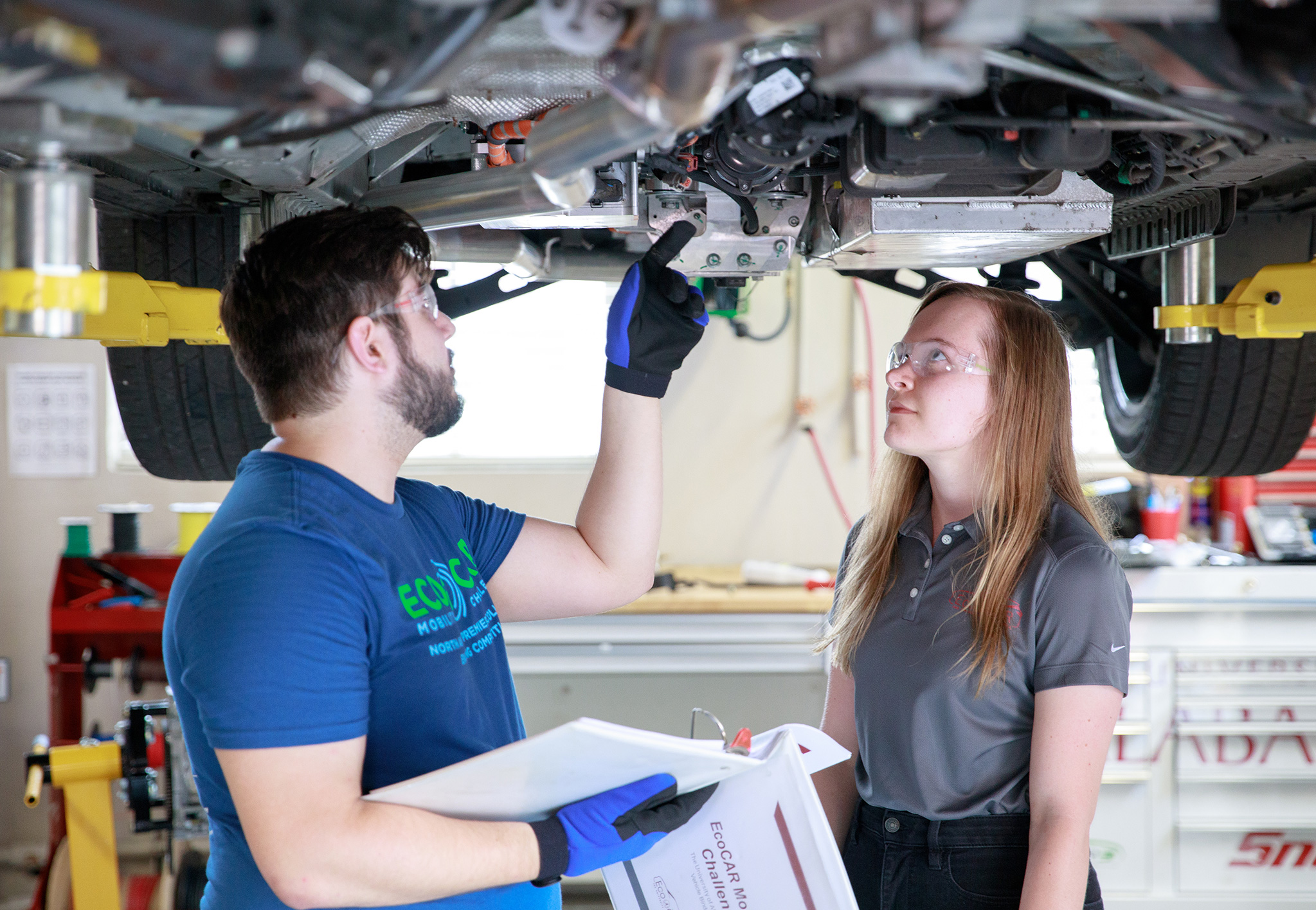Two engineering students in safety glasses examine components on the underside of a vehicle raised on a lift, one holding an EcoCAR binder