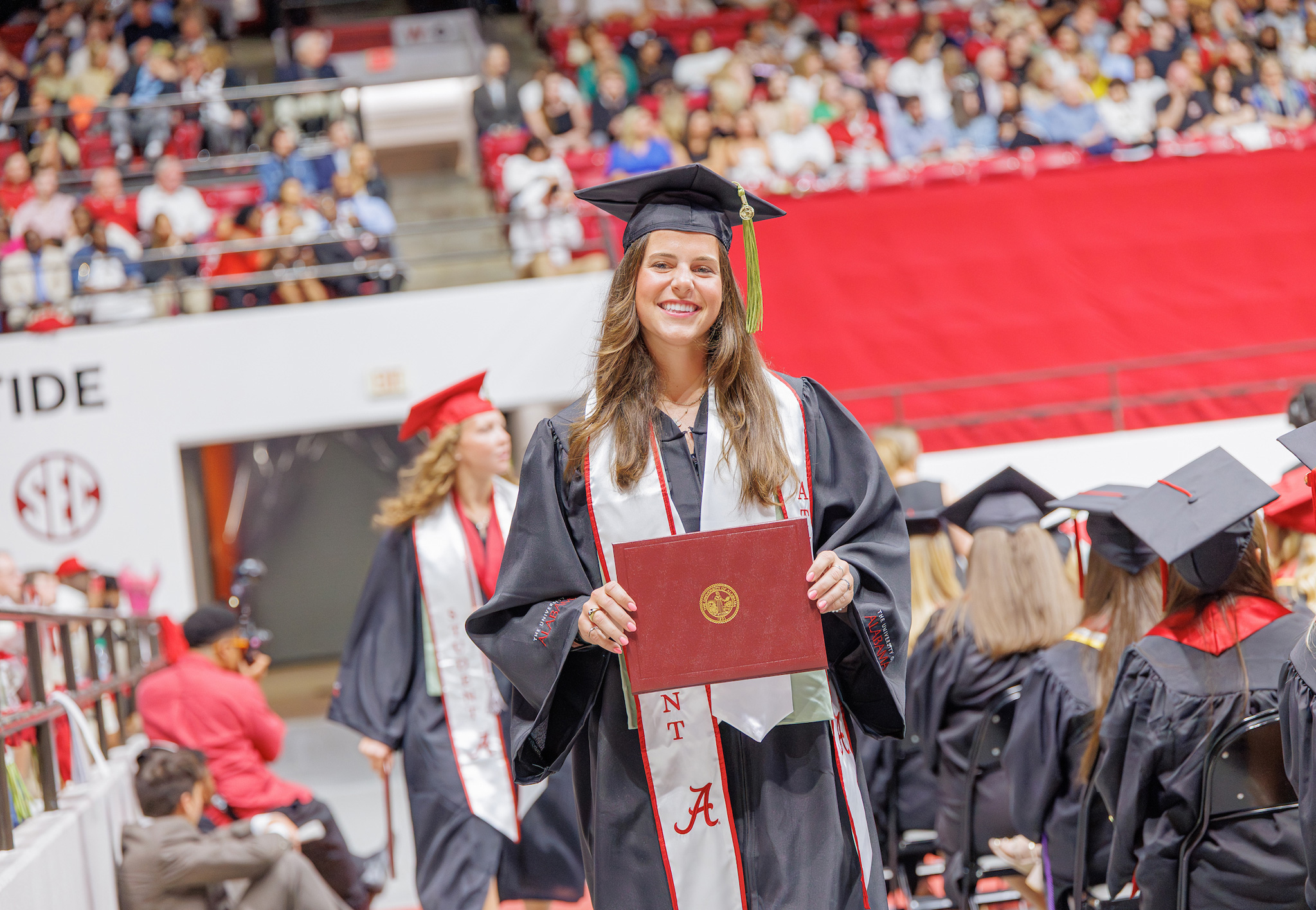 A graduate in cap, gown and honor stoles holds a crimson diploma cover at Coleman Coliseum