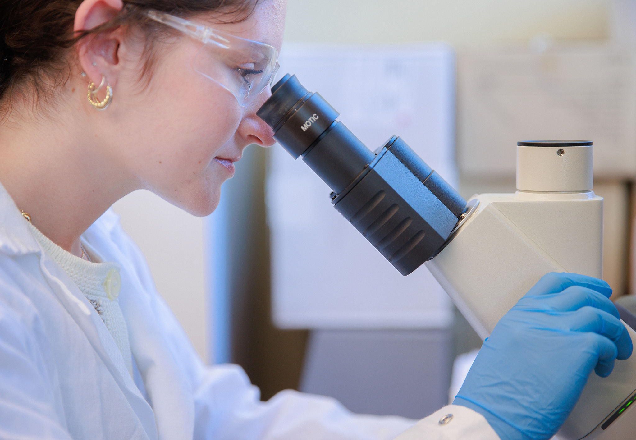 A student in a white lab coat and safety glasses looks through a microscope