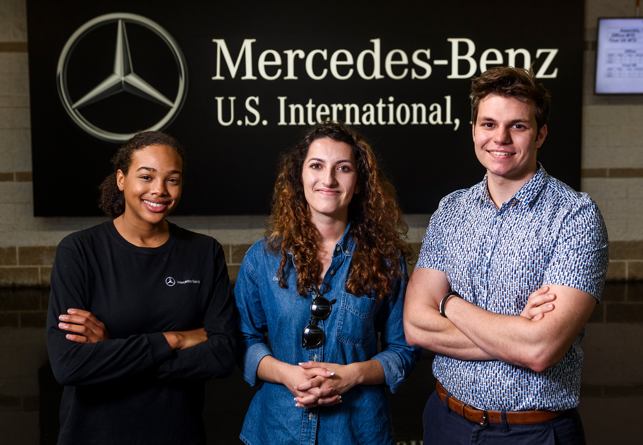 Three students stand in front of a Mercedes-Benz U.S. International sign