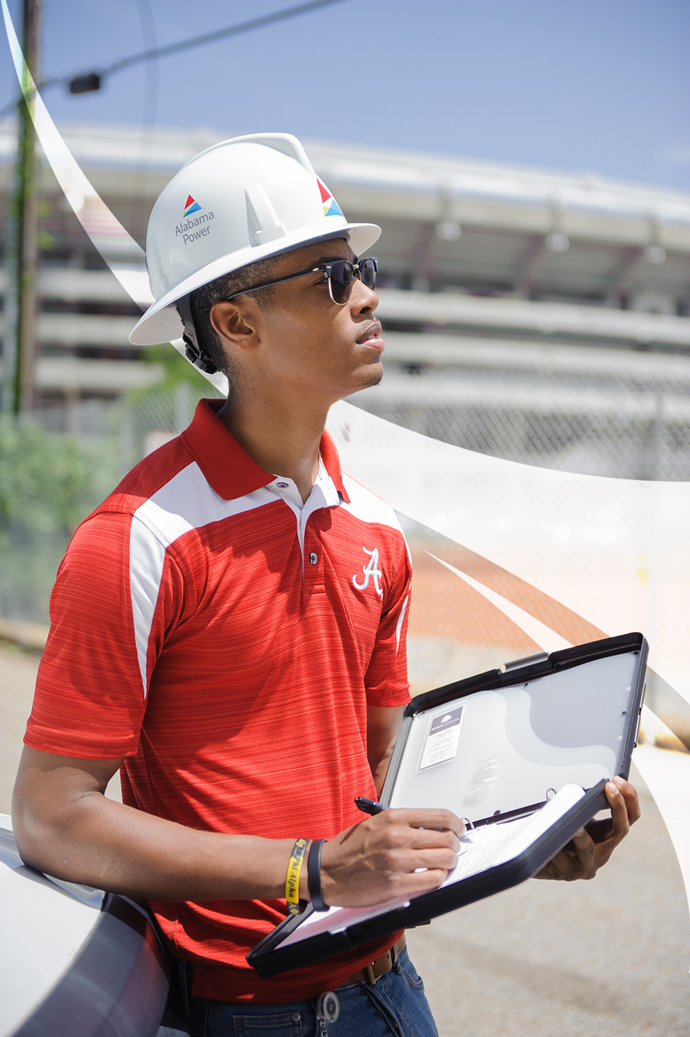A student in an Alabama Power hard hat and crimson polo takes notes on a clipboard outside Bryant-Denny Stadium
