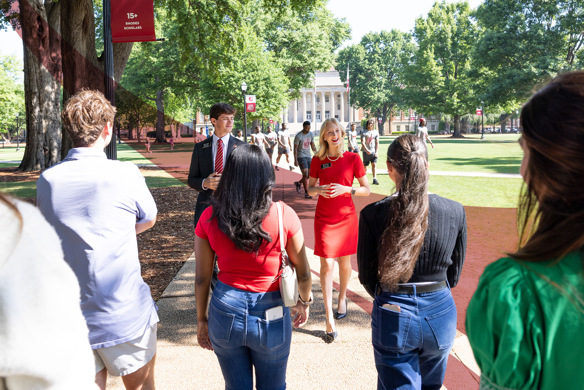 A tour guide in a red dress talks with prospective students on the Quad, Gorgas Library behind her