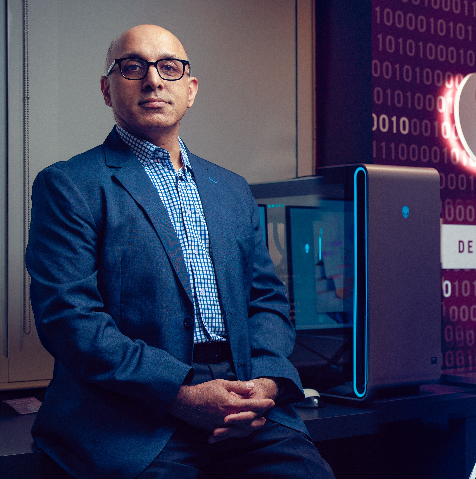Dr. Rahimi seated in a computer science lab, workstations and a lit script A sign behind him