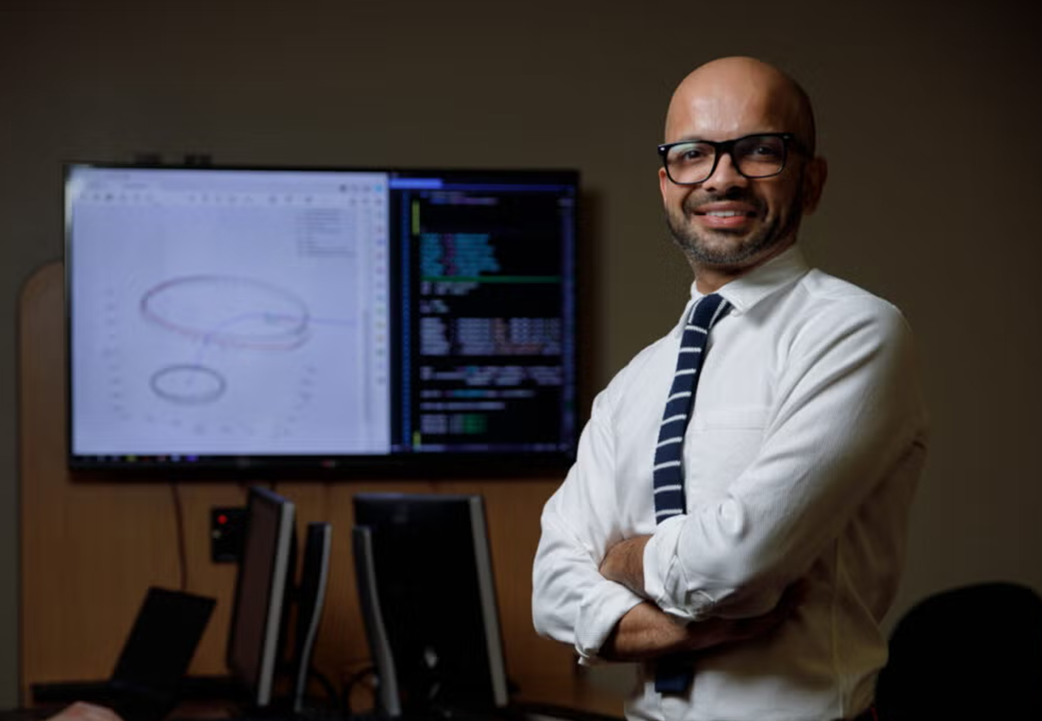 Rohan Sood stands with arms crossed in front of monitors showing an orbital plot and code