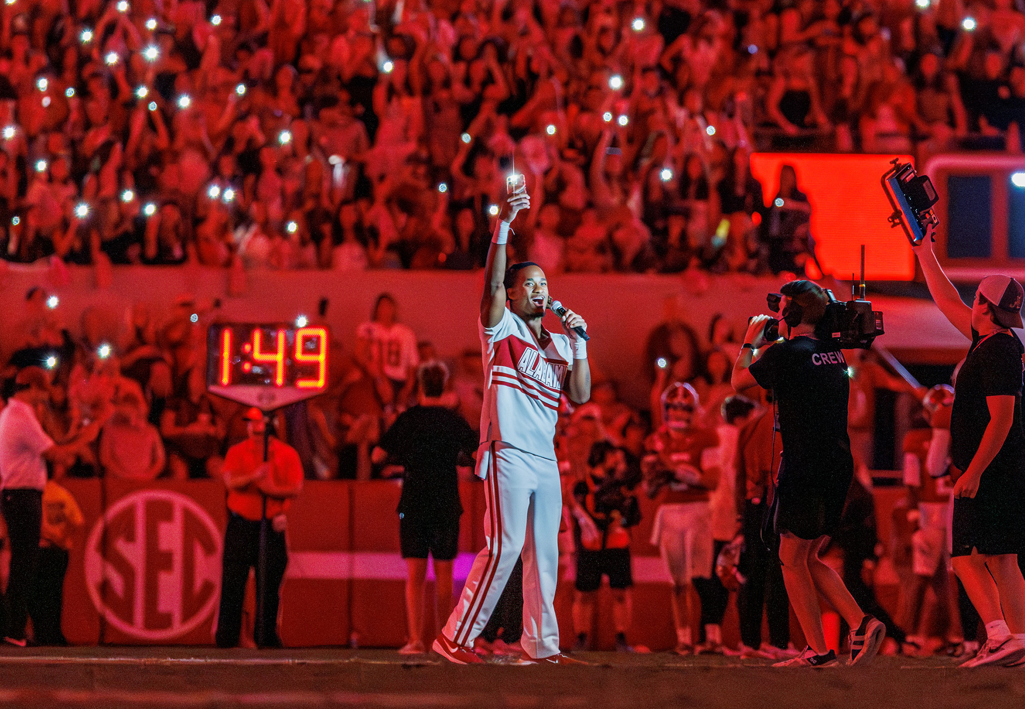 An Alabama cheerleader leads a cheer with a microphone as the crowd raises lit phones behind him